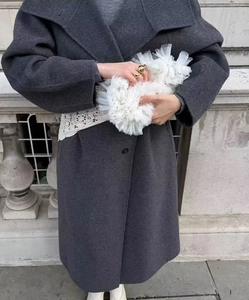 Dettaglio del cappotto lungo in lana grigia con accessorio in pizzo bianco su sfondo urbano