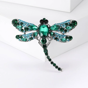 Silver insect dragonfly brooch with green and blue gemstones on a white pedestal