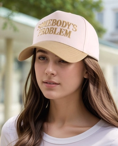 Detail of pink baseball cap with khaki brim and golden 'SOMEBODY'S PROBLEM' embroidery