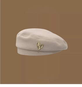 Detail of beige beret featuring a vintage gold heart-shaped metal emblem on textured fabric