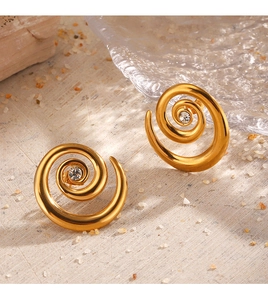 Close-up of golden spiral stud earrings with center crystals on a minimalist sandy surface