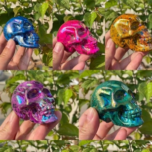 Five assorted aura crystal skulls in rainbow colors shown held against green foliage