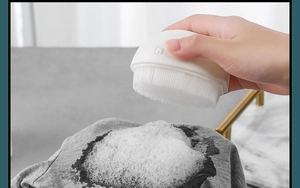Close-up of whale-shaped laundry brush generating white foam on grey fabric surface