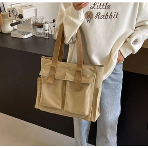 Beige canvas tote bag with front pockets and brown handles held in a lifestyle kitchen setting