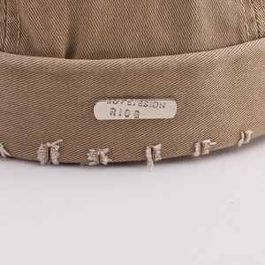 Detail of khaki docker hat showing distressed edges and a silver metal logo plate
