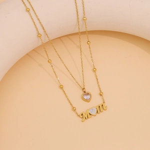 Detail of a double-layer gold necklace with heart pendant and MOM charm on a soft background