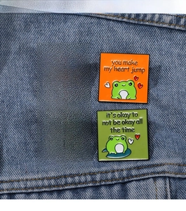 Close-up of four cute frog enamel pins with motivational quotes on a denim jacket