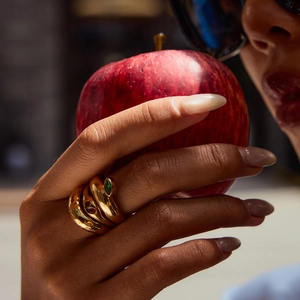 Detail of gold wrap ring with red and green gemstones worn by a model holding an apple