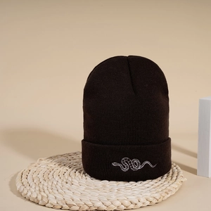 Detailed view of black knitted beanie featuring white snake embroidery on a straw mat