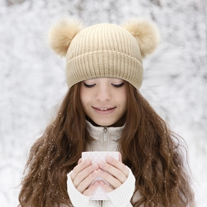 Close-up of beige cable knit winter hat featuring dual faux fur pom-poms on a snow scene