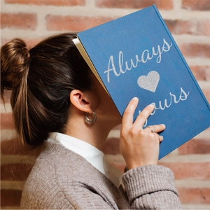 Close-up of blue notebook with silver glitter Always Yours text held against a brick wall