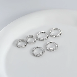 Detalle de pendientes de aro plateados con pedrería y texturas de cadena en base blanca