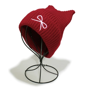 Macro detail of wine red cat ear knit hat featuring a white bow embroidery on metal stand