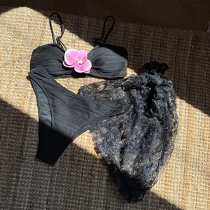Detailed flat lay of a black three-piece bikini set with sheer lace cover-up on a straw mat