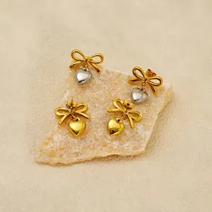 Golden bow stud earrings with heart charms displayed on a minimalist stone backdrop