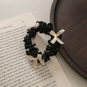 Dettaglio di uno scrunchie nero plissettato con fiocchi color crema su vassoio di legno