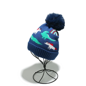 Detail of blue knitted winter hat featuring cartoon dinosaur, unicorn and colorful pom-pom