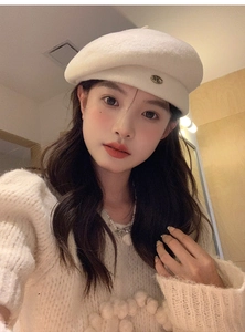 Close-up of a cream-white wool beret featuring a metallic logo on a model in knitwear