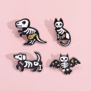 Set of four spooky animal skeleton enamel pins including T-Rex and cat on pink background