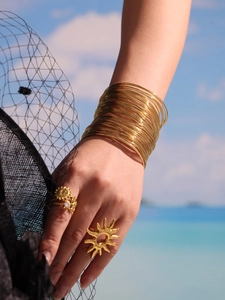 Detail of layered gold cuff bracelet and sunburst ring worn against beach background