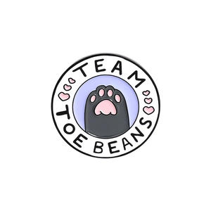 Detail of round metal enamel pin featuring a cat paw design and Team Toe Beans text