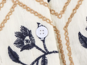 Detailaufnahme blauer Blumenstickerei und weißem Knopf auf cremefarbenem Baumwollleinen