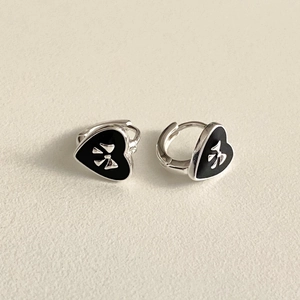 Close-up of heart-shaped silver earrings with black enamel and bow detail on textured paper
