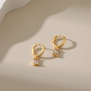 Close-up of gold-plated hoop earrings with rectangular zircon pendants on white stand