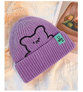 Detail of purple knit beanie featuring bear embroidery and green textile brand label