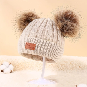 Close-up of beige cable knit winter hat featuring two large faux fur pom-poms and leather tag