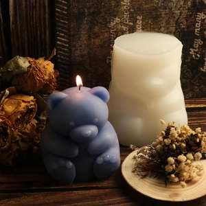 Close-up of a lit blue teddy bear candle next to its white silicone mold on wood