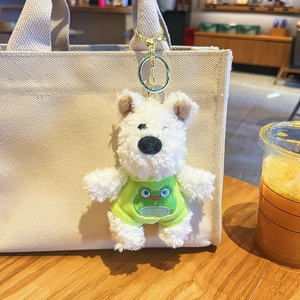 Cute white plush dog keychain in a green frog outfit hanging on a beige canvas bag