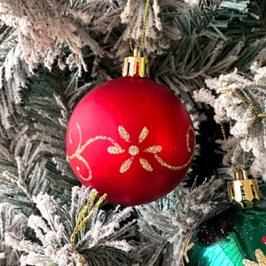 Detail of red Christmas ornament with gold glitter patterns hanging on snowy pine tree branch