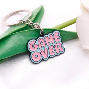 Close-up of a pink Game Over pixel art keychain on a silver ring with plant decor