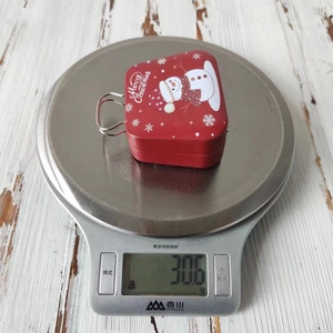 Detailed view of 30.6g Christmas tin box weight on a digital scale with snowman graphics