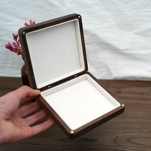 Hand holding an open square wooden jewelry box with white velvet lining and metal hinges
