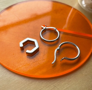 Flat lay of silver geometric hoop earrings and hexagonal ear cuff on orange acrylic disc