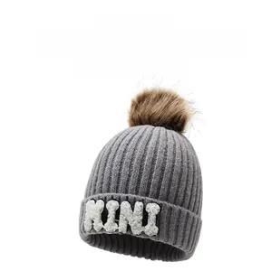 Close-up of gray knit winter hat with brown pom pom and white MINI embroidered letter