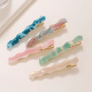 Close-up of five colorful wavy acrylic hair clips with gold metal backings on beige surface
