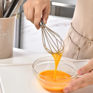 Manual silicone whisk with stainless steel handle stirring eggs in a glass bowl