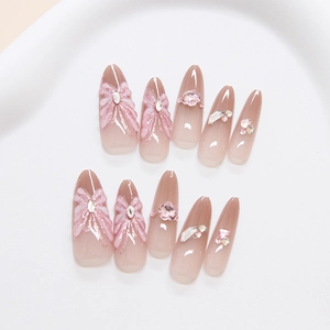 Faux ongles amande dégradé rose avec nœuds 3D et strass posés sur plateau blanc