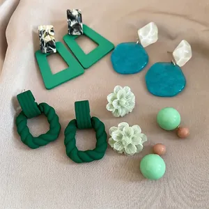 Five pairs of acrylic geometric drop earrings in green tones on beige fabric background