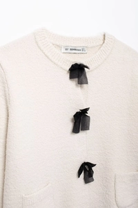 Detail view of the cream knitted cardigan neckline with black ribbon bows and garment label