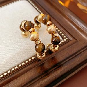 Detalle de pendientes en forma de C con ojo de tigre y cuentas doradas sobre fondo beige