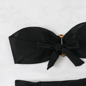 Flat lay of black bandeau bikini set with gold metal ring and bow detail on marble background