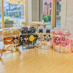 Dual-compartment cartoon water bottle with straw and strap in a sunny cafe setting