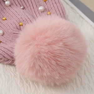Pink pearl-studded knit hat with large faux fur pom-pom on soft white textured background