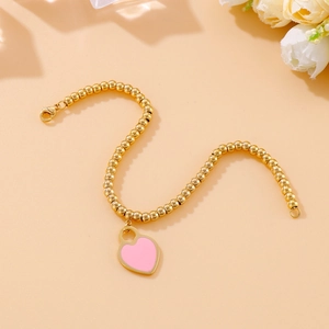 Detailed view of pink heart pendant and 18k gold plated beaded bracelet on peach surface