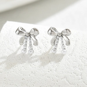 Silver bow stud earrings with dangling white pearl accents on elegant stone background