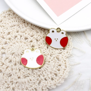 Detailed view of two white enamel frog charms with red cheeks and gold-toned metal plating
